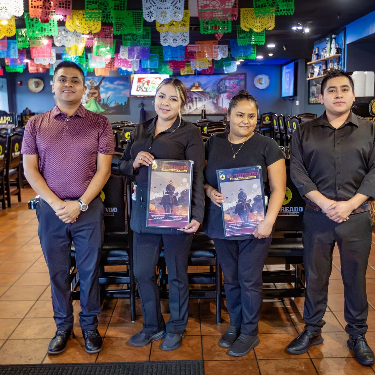 The El Toreado team standing in the West Des Moines dining room under colorful papel picado, holding menus